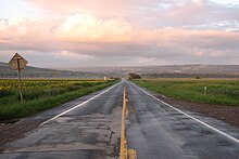 A two-lane paved road, wet in spots, stretching straight ahead with fields on either side towards a ridge in the distance. Above it in the sky is a cloud with orange-tinted sunlight on it; there is also a low mist over the fields in the distance.