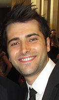 A man dark hair wearing, black suit, including black tie and white shirt.