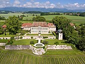 Photo couleur du château de Vuillerens prise en 2016 depuis les airs. On y voit la cour d'entrée principale et le devant du bâtiment en contrebas, ainsi que les jardins et les fontaines de la cour principale.