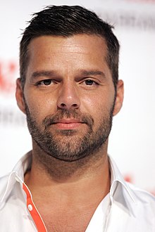 A bearded man, facing forward, wearing a white shirt with an orange strip.