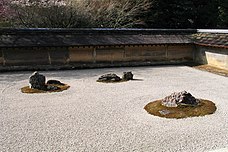 Um jardim tradicional japonês coberto de areia, com três pedras maiores posicionadas de forma equidistantes. O jardim é envolto por um muro baixo com um pequeno telhado.