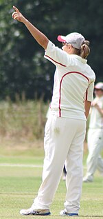 A woman with light brown hair wearing white cricket uniform with red piping points with her left arm. Another player is visible in the background.