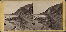 Two men standing on the Hudson River shore with railroad tracks paralleling the shoreline and a mountain off in the distance