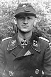A black-and-white photograph of a man wearing a military uniform, cap and a neck order in shape of an Iron Cross. His cap has an emblem in shape of a human skull and crossed bones.