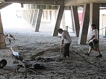 Two men, dressed in white shirts and loose pants, are seen beneath a wooden pavilion. The pavilion's legs stick out diagonally from the pavilion. One man carries a pipe which he points toward a pit in the sandy ground. The second man walks toward him.