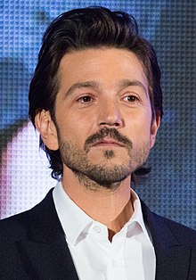 A man (Diego Luna) with well groomed dark hair and a beard wearing a suit.