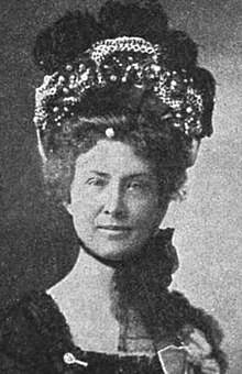 A white woman wearing a large, elaborate bonnet, tied under her chin with a mass of tulle.