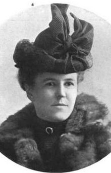 A young white woman wearing a fur coat, a dark high-necked blouse, and a large dark hat.