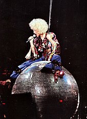 Image shows female performer sitting on a disco ball, wearing a big curly blonde wig, a maroon blouse and blue pants with red heels and is holding a microphone to her mouth.