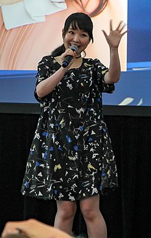 Itō in a black dress holding a microphone in one hand and waving with the other