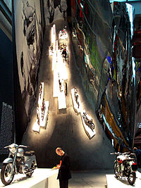 A man standing between two vintage motorcycles on pedestals surrounded by curving, reflective walls.