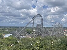 An overview of Iron Gwazi in 2023. The roller coaster sits center in the image with most of its elements visible. Foliage lines the foreground at the bottom of the image. The station sits bottom left.