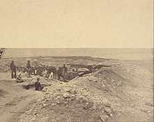 Photographie d'une position d'artillerie au sommet d'une colline rocailleuse et désolée où sont déployés deux canons derrière des talus renforcés de sacs de sable. Plusieurs dizaines d'hommes portant des tenues variées prennent la pose.