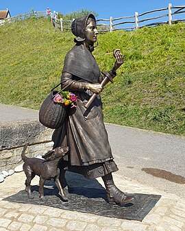 A bronze statue of Mary Anning. She is walking, holding an ammonite with a basket over one arm and her dog trailing behind.