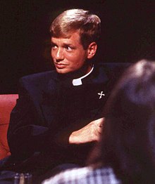 A man wearing a cassock and Roman collar, with a pin badge of the Dominican Order's coat of arms on his left lapel