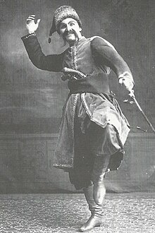 A man in a dance pose, one knee raised, one arm overhead, wearing costume and a long mustache