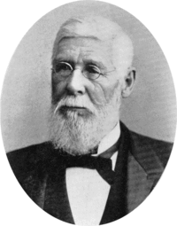 A black-and-white photograph of a man with white hair, mustache and beard wearing glasses