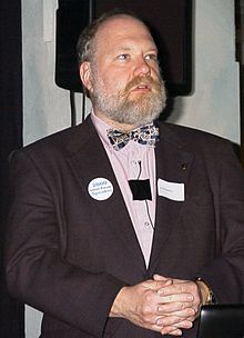 Balding white male with grey beard, wearing a black suitcoat, pink shirt, and patterned bow tie, standing with hands clasped.