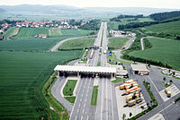 Aerial view of a four-lane motorway crossing green fields, with a small village with a church spire in the distance to the left of the motorway. In the foreground, there is a white roof structure, resting on slim white pillars, across all four lanes of the motorway; to the left, the roof also extends over a slip lane which branches off from the main road and then rejoins it; on the right, just before the roof structure, there is a parking lot with diagonally parked orange and brown lorries.