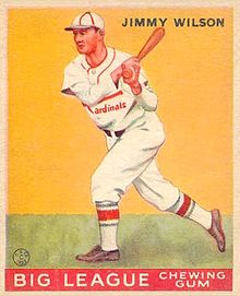 A baseball card image of a man wearing a white baseball uniform and cap with red trim swinging a baseball bat