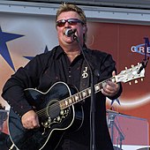 A man wearing dark glasses and a black shirt playing a guitar and singing into a microphone