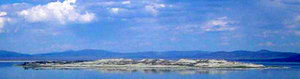 A small, dark island rises above the calm, blue surface of a mountain-rimmed lake.