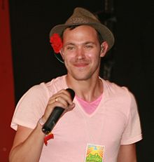 A man in a pink t-shirt smiling and holding a microphone