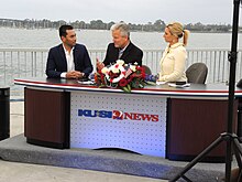 Three people at a news desk with a "KUSI 9 News" logo positioned with a waterfront backdrop