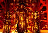 Front view of three standing statues which are covered in gold. The central statue is about two heads taller than the two falnking statues. It has the right hand pointing down with the palm towards the viewer and the palm of the left hand turned upward with the thumb touching the middle figer. All three statues have halos behind their heads with emanating rays. Color photograph.