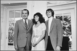 A dark-haired older man in a suit, a young woman with long dark hair wearing a dress and a young man with mid-length hair wearing a jacket over a rollneck sweater