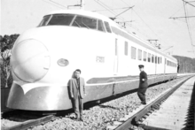 Schwarz-Weiß-Aufnahme vom zweiteiligen Zug A der JNR-Baureihe 1000 aus dem Jahr 1963. Der Triebzug steht auf freier Strecke und zwei Bahnangestellte, einer in Anzug und einer uniformiert, stehen vor dem vorne sichtbaren Endwagen A-021, während ein dritter Angestellter sich bei der Führerstandstür des anderen Endwagens befindet.