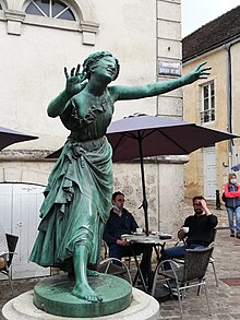 Statue représentant une femme aux yeux recouverts dans une ville avec des personnes à l'arrière-plan.