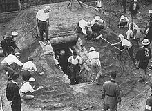 Black and white photo of men and women working on constructing an earthen mound with a doorway cut into it. The doorway is lined with sandbags.