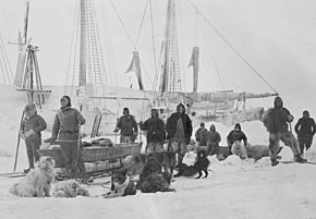 Un groupe d'homme pose aux côtés de chiens et de traineaux, la silhouette d'un navire est visible à l'arrière-plan.