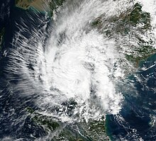 A photograph of a tropical storm near the Malay Peninsula. Although most of the cloudiness and thunderstorms associated with the system are amorphous and disorganized, it is exhibiting healthy outflow on its western flank, as indicated by a large number of thin, relatively straight, cirrus clouds pointing outward from the western half of its circulation.