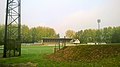 Tribune d'un terrain de football en herbe, derrière un pied de mat d'éclairage et une butte de terre.