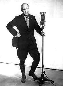 Full body photograph of DeMille wearing a blakc suit, holding a top hat in one hand and the CBS radio microphone in the other