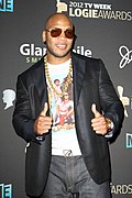 An Afro-American male donning a black peacoat, ash-coloured jeans, dark sunglasses, a white shirt with scarlet lettering, and a gold chain about the throat, gives the thumbs-up gesture on backhands while smiling in front of a gray background
