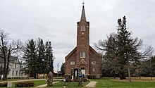 brown brick church with a tall steeple and a statue of the Holy Mary in front, flanked by pine trees