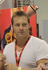 A mid-aged man with short brown hair, is sitting behind a table with a pen in his hand, and is looking to his left.