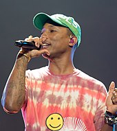 An image of Pharrell Williams holding a microphone in his hand, wearing a red T-shirt along with a green cap.