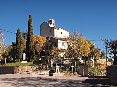 Place Fuata - de gauche à droite : le monument aux morts entouré de ses deux Cyprès , l'église sur son piton rocheux, la fontaine et l'abri-bus scolaire.