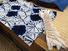 A rectangle of indigo blue and white cloth lying on a wooden table covered with a tatami mat, viewed diagonally from the corner.