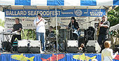 Four people on a stage; from left to right, they are playing a keyboard, a trumpet, a drum set, a guitar, and a saxophone. In the background are various signs, and on the stage are microphone stands and other audio equipment.