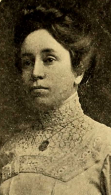 A 1911 yearbook photograph of a young woman with dark hair in a bouffant updo, wearing a high-collared white lacy shirtwaist or dress, and a pendant