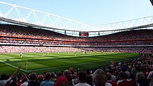 Vue depuis la tribune basse d'un grand stade avec ses tribunes remplies avec un match en cours.