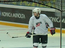 An ice hockey player on the ice. He is wearing a white jersey with the letters "USA" on the front in red and blue.