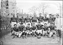 Photographie noir et blanc d'un groupe d'hommes. Au premier plan sept hommes en maillot et short sont à genou ou en position accroupie. Devant le cinquième est posé au sol un ballon ovale. Un homme vêtu d'une veste et d'un pantalon se tient debout à droite de l'image. Derrière ces huit hommes, dix hommes sont debout dont huit sont en tenue de sport. Derrière les personnes à gauche de l'image se trouve un immeuble de plusieurs étages. Quelques arbres poussent derrière au loin.