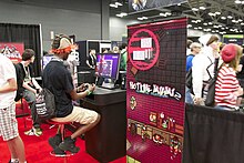 A man with red hair, a black shirt, and cargo shorts plays a demo of Hotline Miami 2 with a video game controller. Several other people are in the background, and promotional material for the game sits next to him.