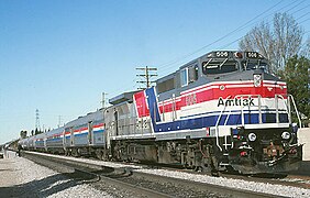 A gray diesel locomotive with red, white, and blue stripes wrapping around the sides and front. There is one wide stripe of each color, plus several narrower stripes. The stripes angle upwards and cross each other on the side of the locomotive
